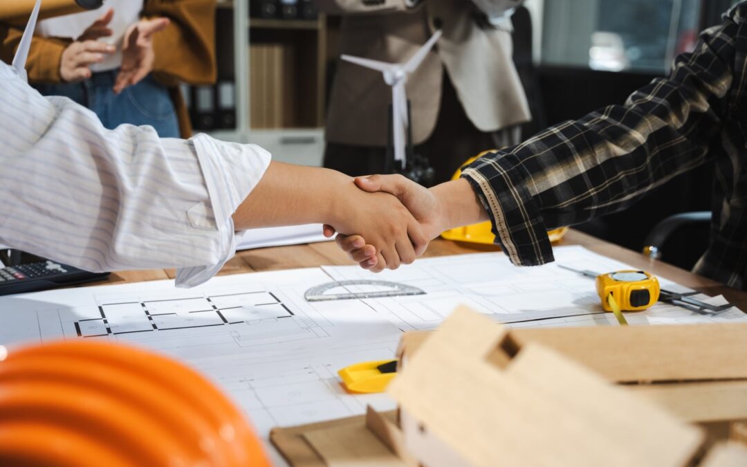 Two people shake hands over architectural blueprints spread on a worktable, with a tape measure and hard hat nearby in a construction setting.